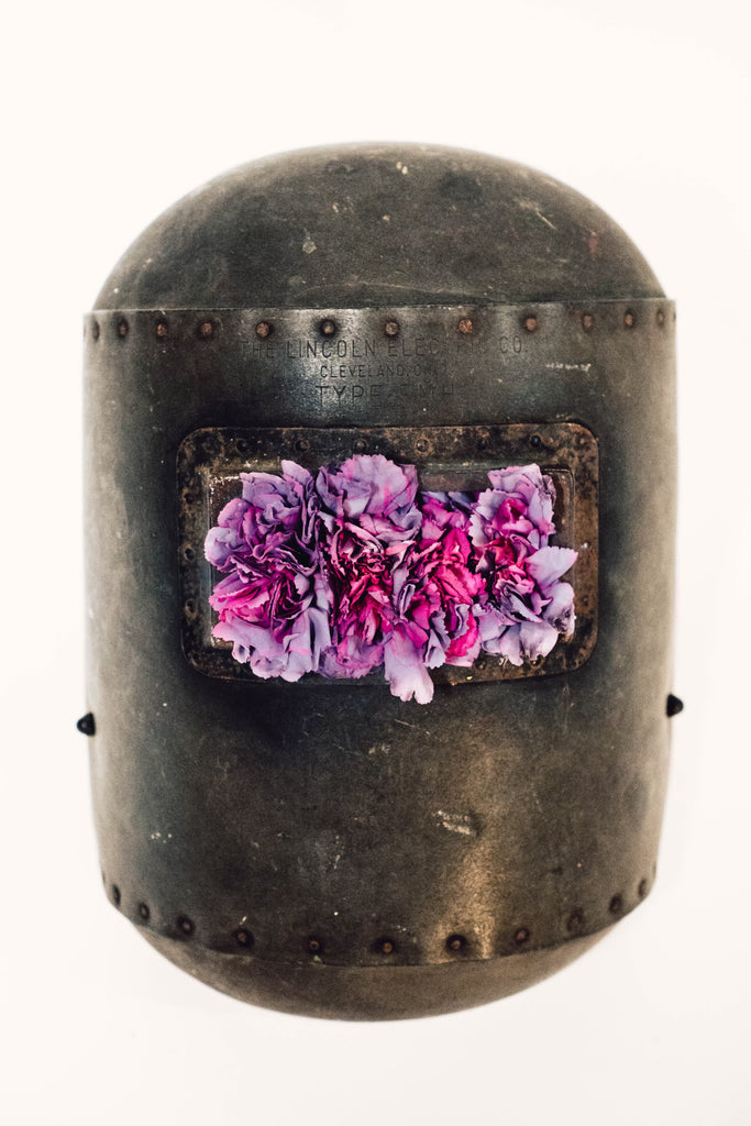 photograph of welder's mask filled with purple flowers where eyes should be