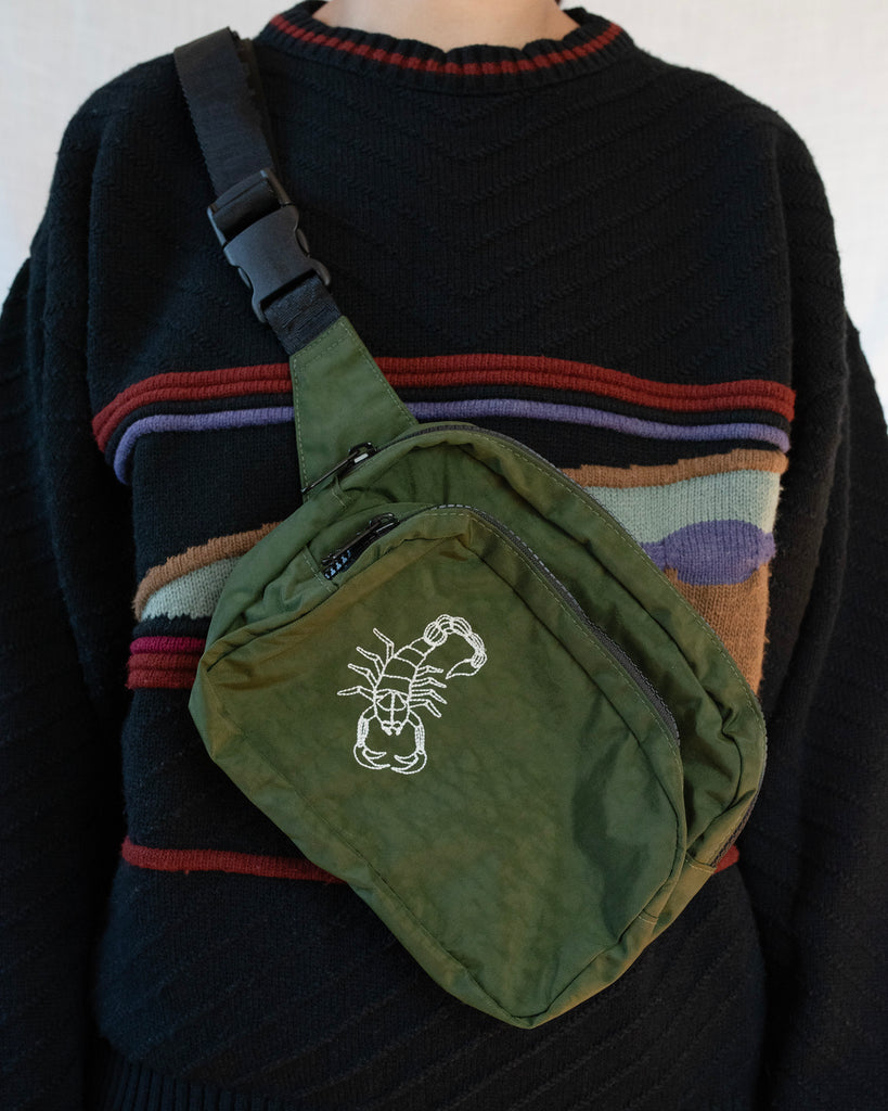 dark green fanny pack with white embroidered scorpion worn by a person in a sweater